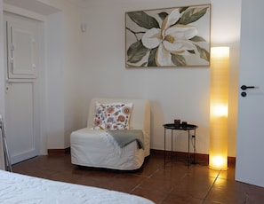 Ein Zimmer mit einem Bett, einem Sessel mit floralem Kissen, einem Blumenbild, einer Stehlampe und einer Tür.