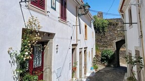 A narrow cobblestone street flanked by white buildings with red shutters and a stone bridge in the background. - Encanto da Aldeia (Castelo de Vide)