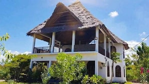 A two-story house with a thatched roof, a balcony on the upper floor, and a garden in the foreground. - Villa Noemi by the Sea (Bwejuu)
