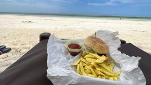A beachside meal setup with a burger and fries on a black mat, a small container of ketchup, and a pair of flip-flops on the sand. - Villa Noemi by the Sea (Bwejuu)