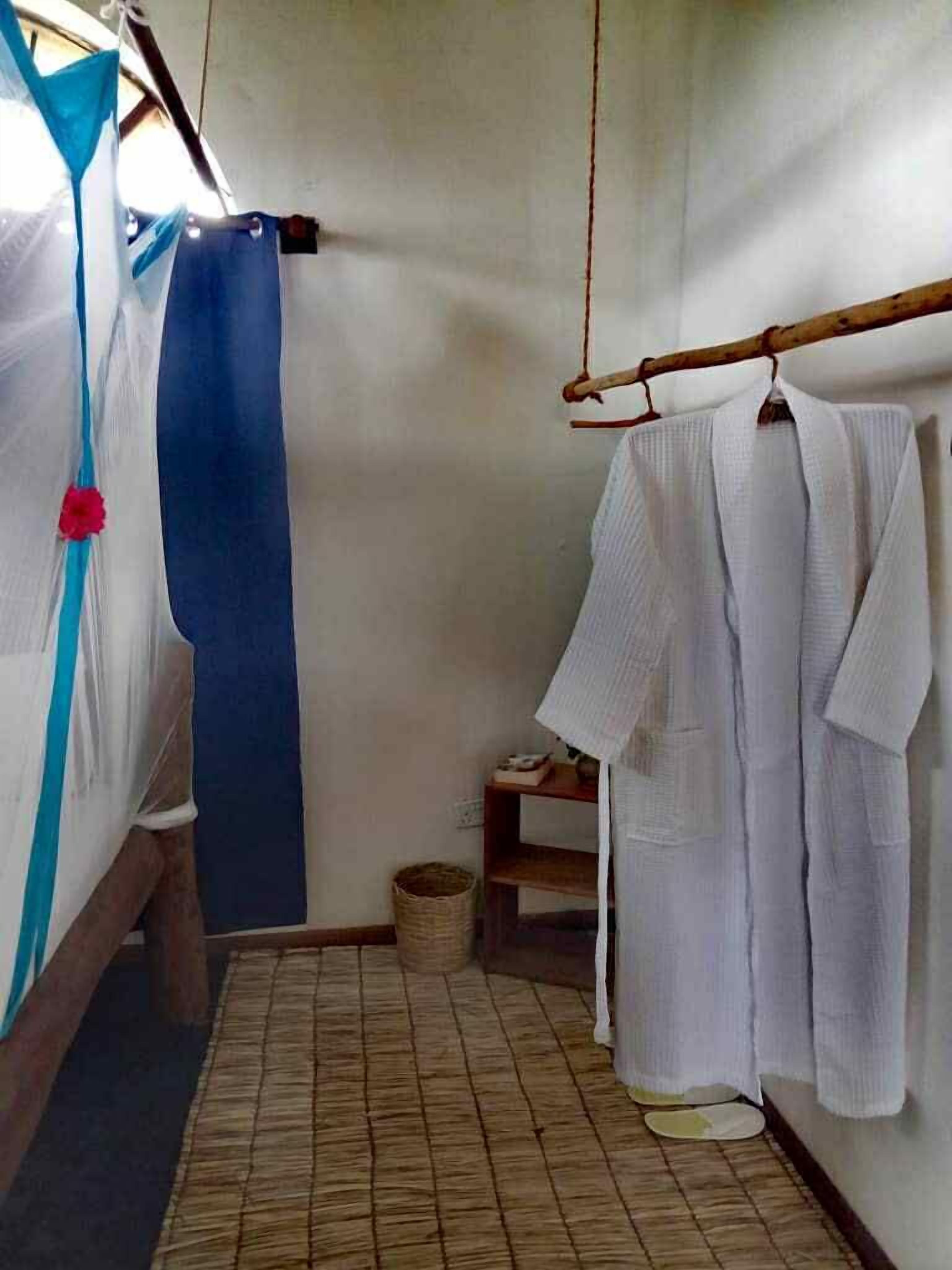 A small, narrow room with a tiled floor, a wooden bench, a hanging rack with white robes, and a blue curtain.