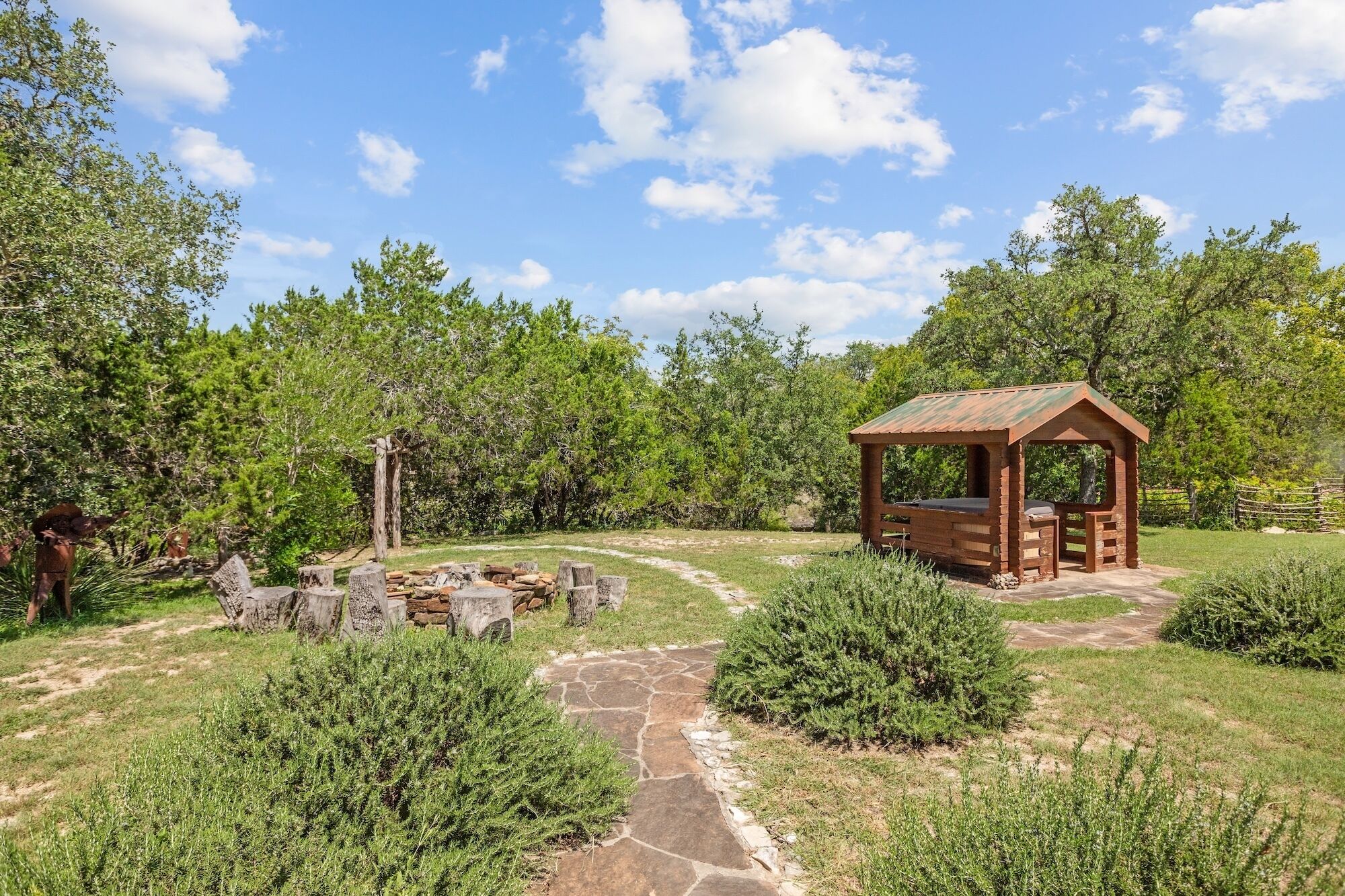 A wooden gazebo with a stone pathway leading to it, surrounded by greenery and trees.
