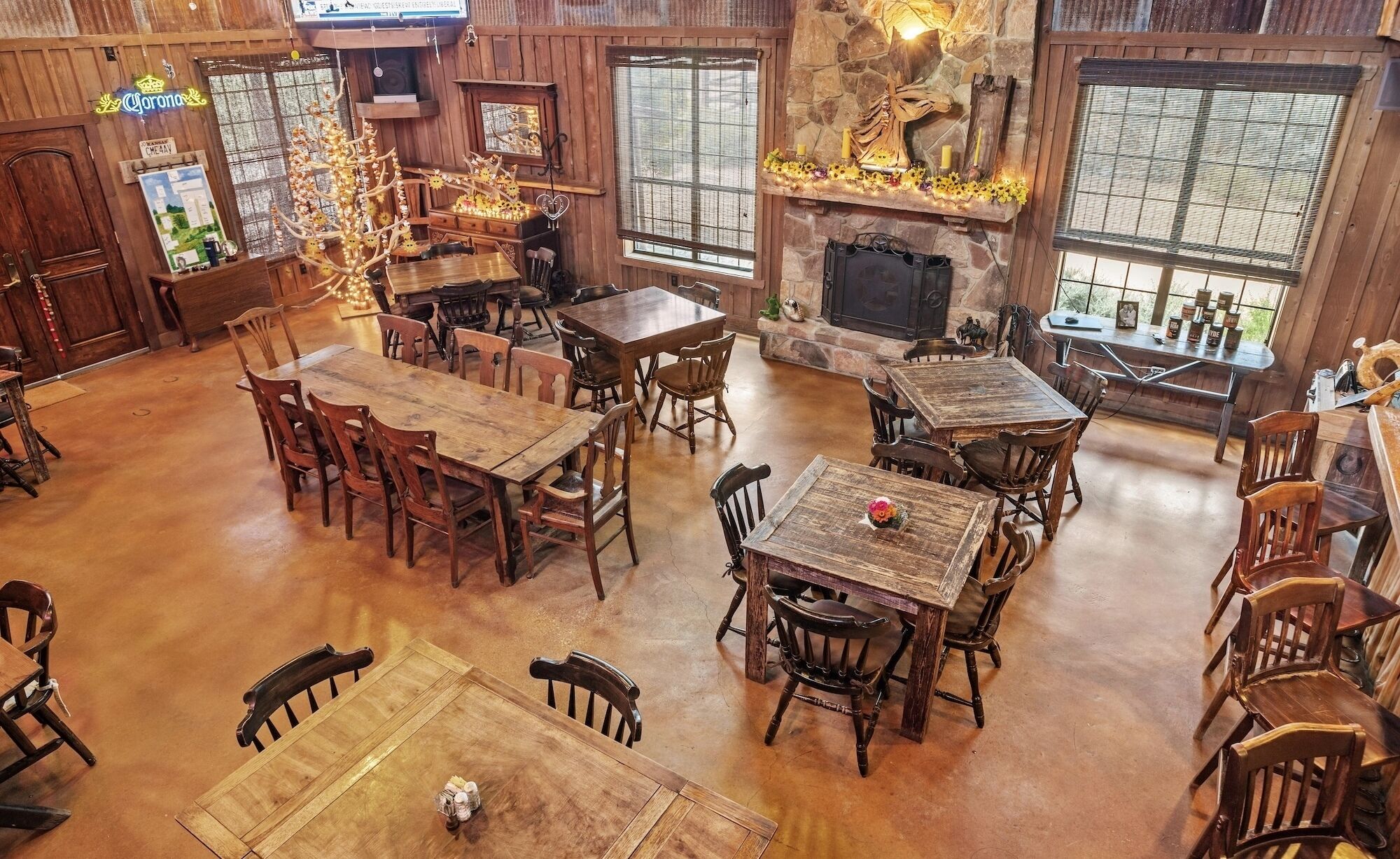 A rustic interior with multiple wooden tables and chairs, a stone fireplace, and large windows.