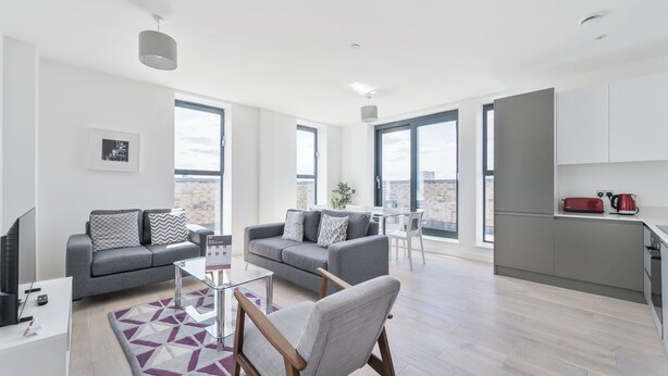 A modern living room with a grey sofa, a wooden chair, and a glass coffee table. There is a kitchen area with a red kettle and a white countertop. - Roomspace Apartments -Caroline Street (London)