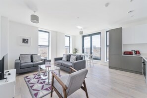 Uma sala de estar moderna com um sofá cinzento, uma cadeira de madeira e uma mesa de centro de vidro. Há uma área de cozinha com uma chaleira vermelha e uma bancada branca.