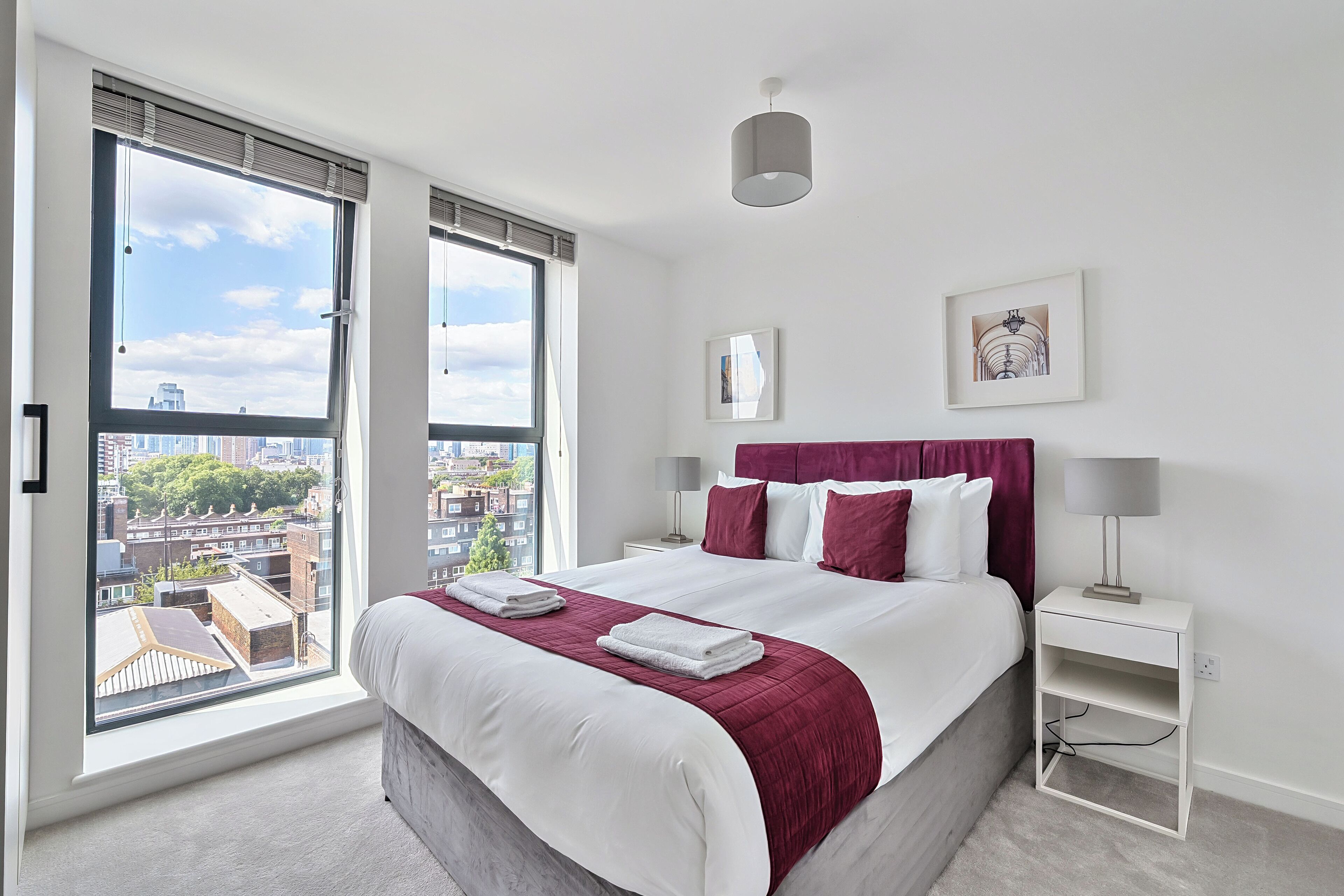 Una habitación de hotel moderna con una cama grande, ropa de cama blanca, una manta roja y vistas a la ciudad a través de amplios ventanales.