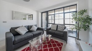 A modern living room with two grey sofas, a glass coffee table, and a red rug. There is a large window with a view of a balcony and a potted plant. - Roomspace Apartments -Caroline Street (London)
