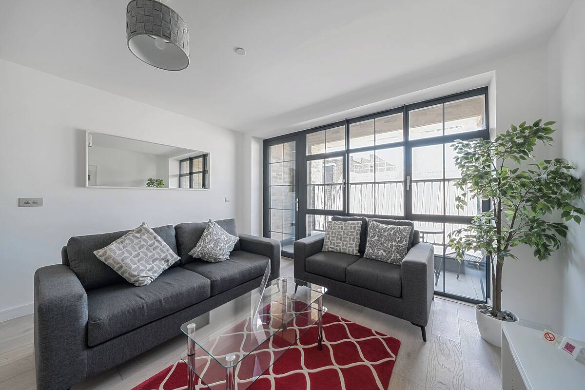 Ein modernes Wohnzimmer mit zwei grauen Sofas, einem Glastisch und einem roten Teppich. Ein großes Fenster bietet Blick auf einen Balkon und einen Blumentopf.