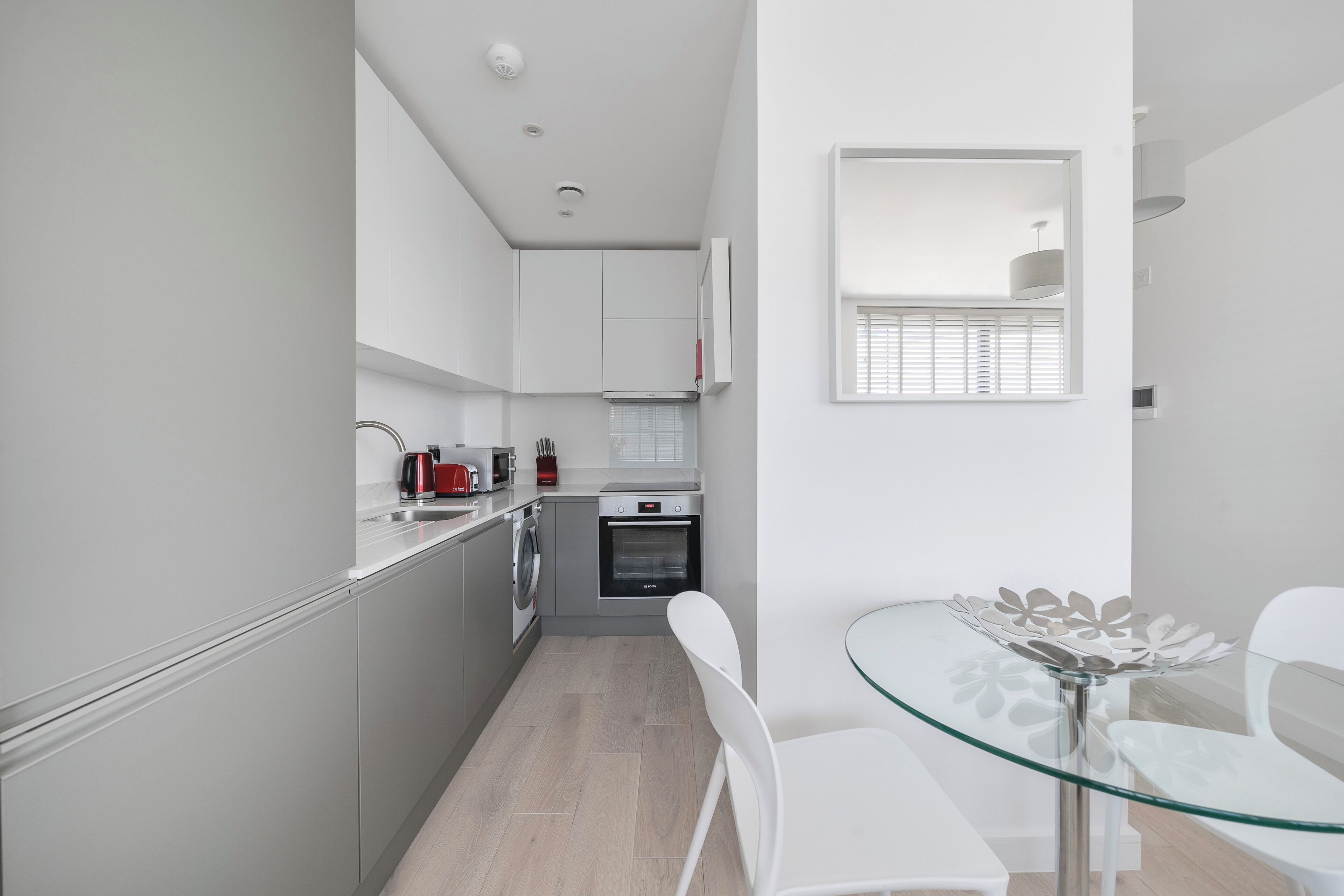 A modern kitchen with white cabinets, a built-in oven, and a glass dining table with chairs.