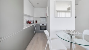 A modern kitchen with white cabinets, a built-in oven, and a glass dining table with chairs. - Roomspace Apartments -Caroline Street (London)