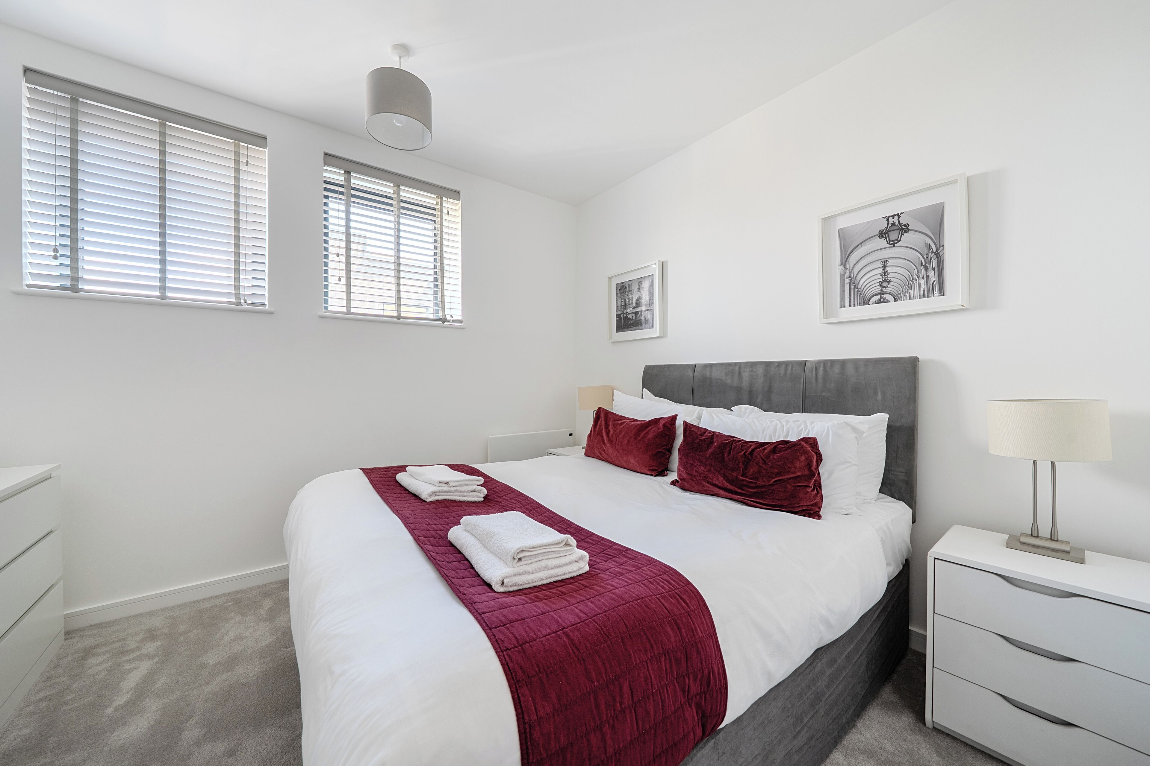 A modern bedroom with a large bed, white walls, and a grey headboard. There are two bedside tables with lamps, a small bedside table with a lamp, and a framed picture on the wall.