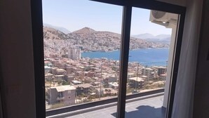 A room with a large sliding glass door leading to a cityscape view, including a body of water and distant mountains. - Brian's Rooms (Sarande)