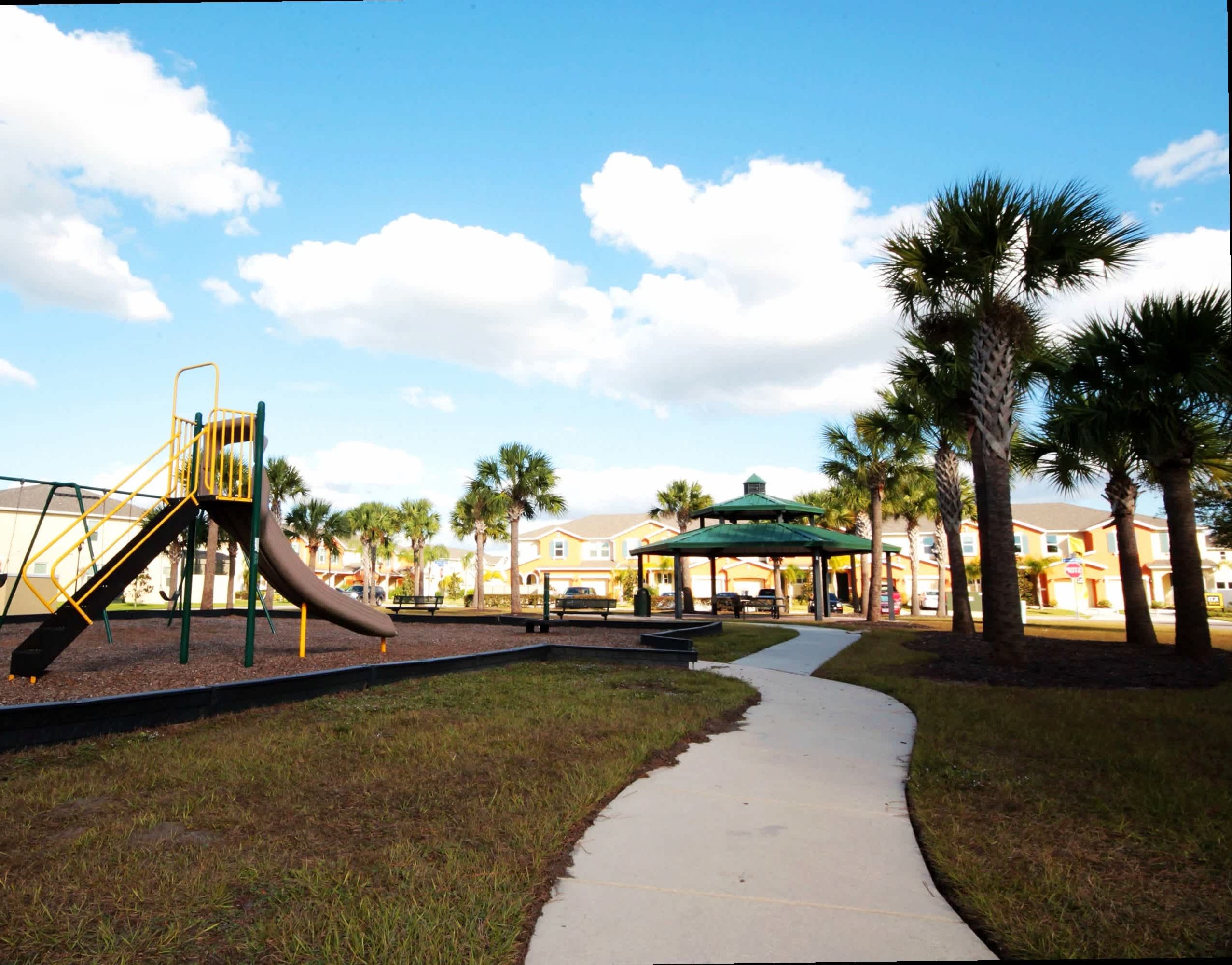 A playground with a slide, surrounded by grass and palm trees, with a pathway leading to a building with a green roof.