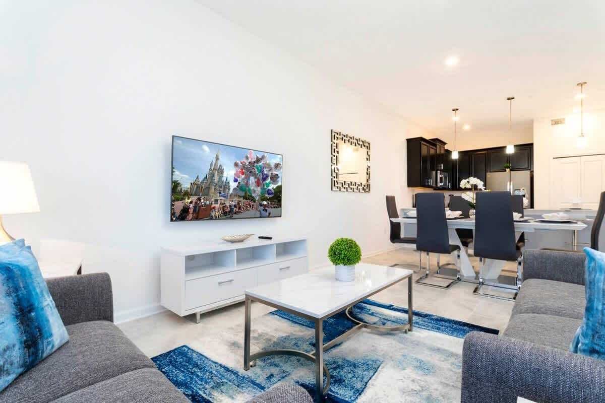 A modern living room with a flat-screen TV, a white coffee table, and a dining area with a black dining set.
