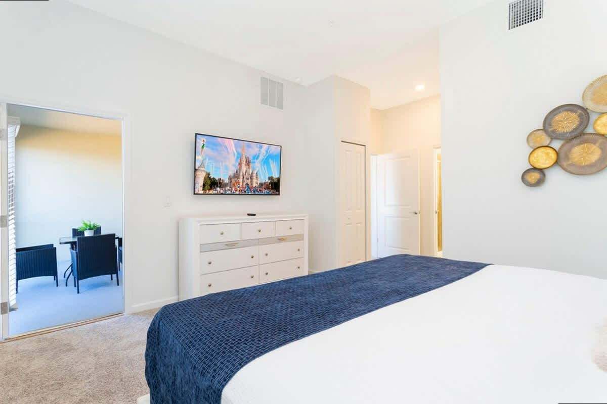 A modern hotel room with a large bed, a flat-screen TV mounted on the wall, a white dresser, and a mirror reflecting a dining area.
