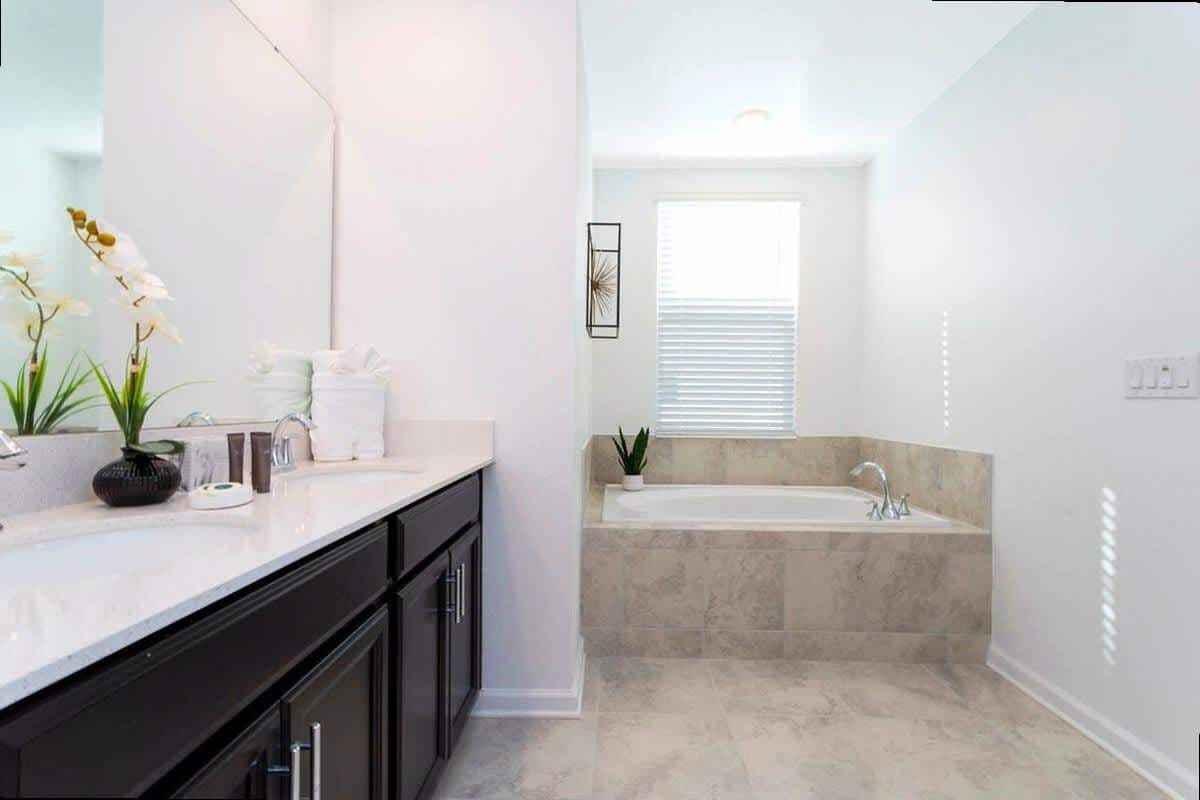 A modern bathroom with a large mirror, a white sink, a black cabinet, a beige bathtub, and a window with blinds.