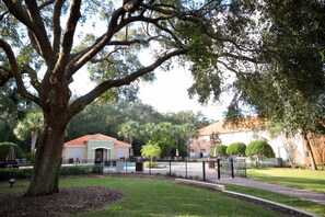 A tree-lined courtyard with a swimming pool, a fenced area, and a building with a red roof. - Modern 4BR w Patio Resort Pool Compass Bay (Kissimmee)