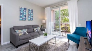 Ein modernes Wohnzimmer mit einem grauen Sofa, einem blauen Sessel und einem weißen Couchtisch. Zwei Wandbilder und eine Schiebetür zu einem Außenbereich sind ebenfalls vorhanden.