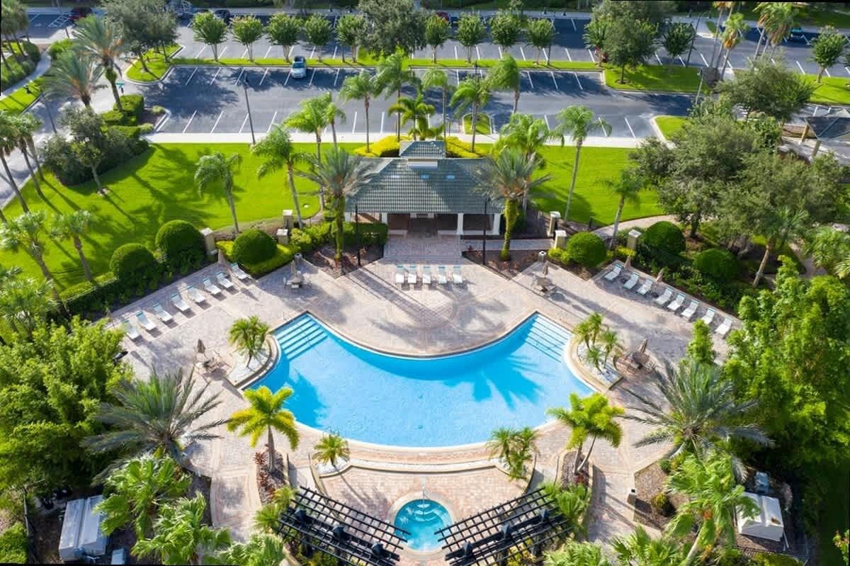 An aerial view of a resort with a central swimming pool, surrounded by palm trees and lounge areas.