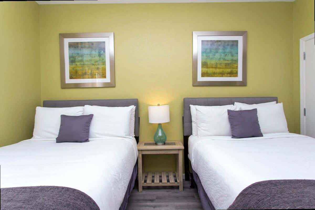 A hotel room with two beds, a small bedside table with a lamp, and two framed pictures on the wall.