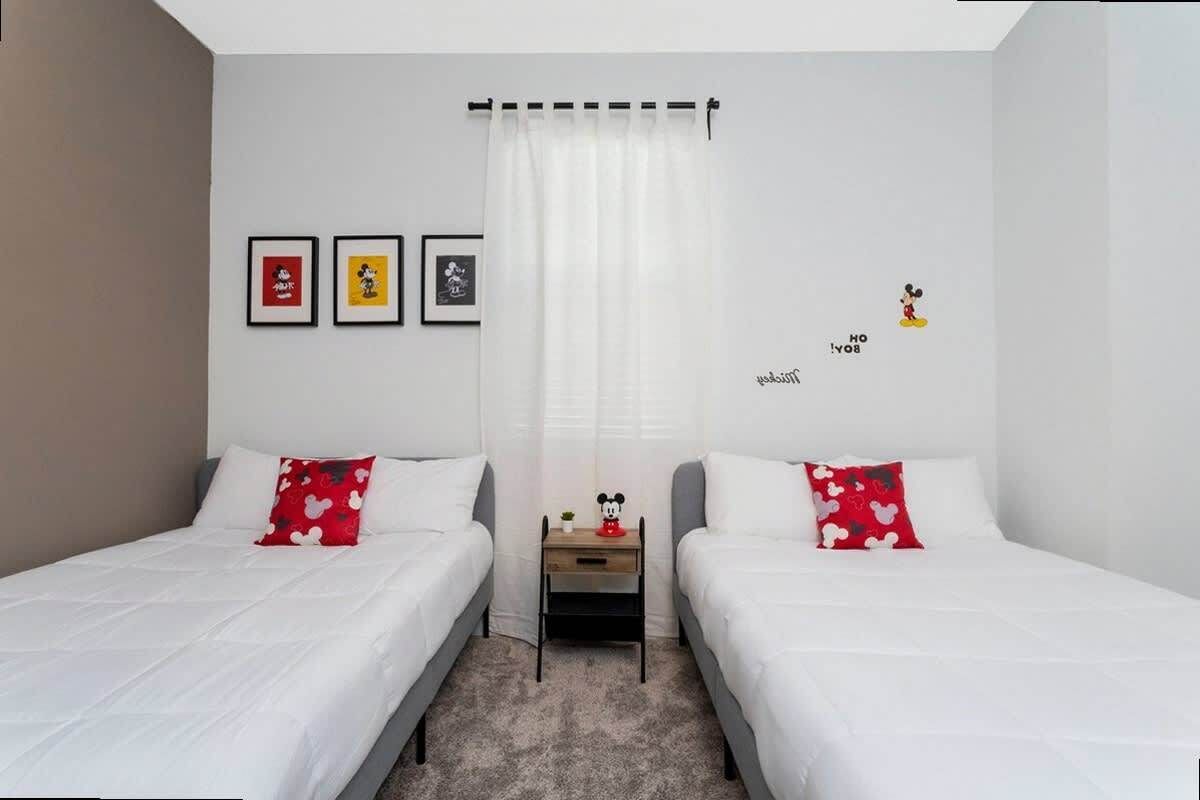 Una habitación con dos camas, una mesita de noche y una ventana con cortinas. Hay cuadros en la pared y un adhesivo de Mickey Mouse.
