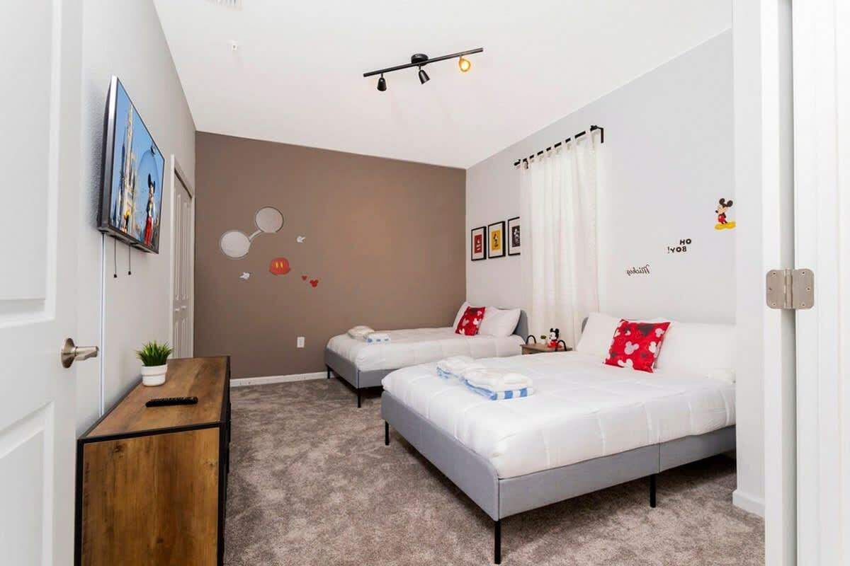 A modern bedroom with two beds, a small wooden side table, a wall-mounted TV, and a ceiling light fixture.