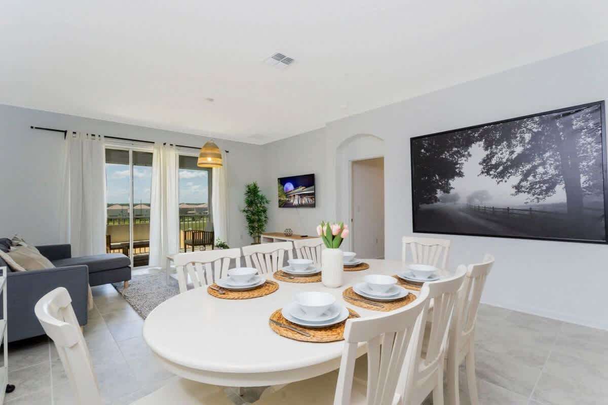 A modern dining area with a round table set for six, white chairs, and a white vase with flowers.