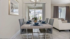 A dining area with a wooden table, chairs, and a chandelier. There is a sofa and a window with curtains in the background. - Bright 4Bd With Pool Resort Perks Near Disney (Kissimmee)