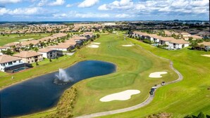 Campo de golf con búnkeres, un estanque y edificios residenciales al fondo.