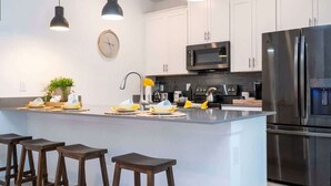 Une cuisine moderne avec des meubles blancs, des appareils en acier inoxydable, un éclairage suspendu et un îlot central doté de tabourets.