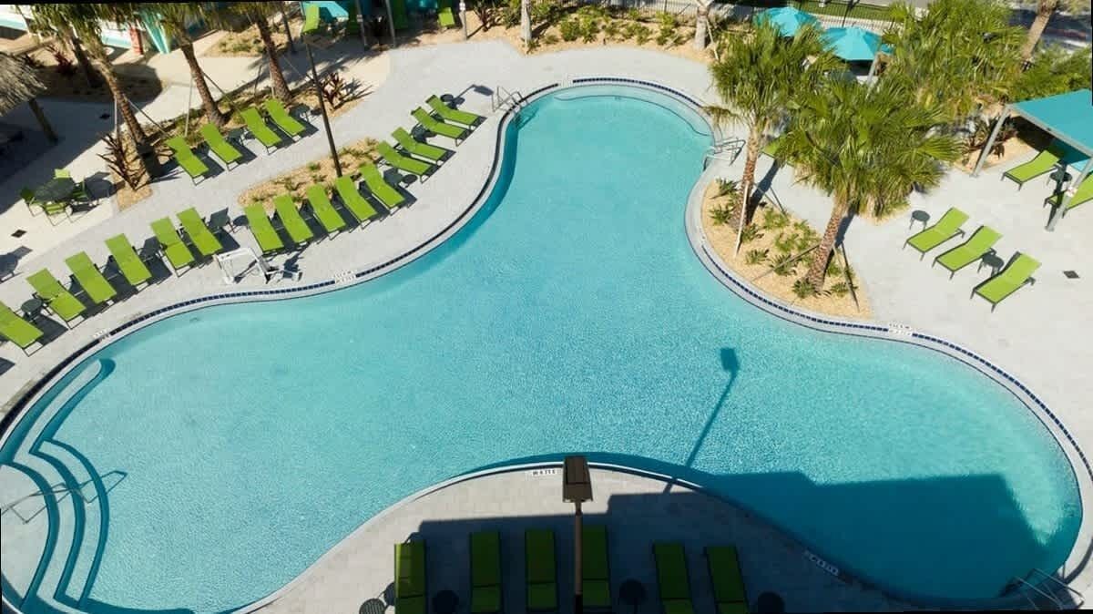 Vue aérienne d’une piscine au design original, avec des formes ondulées, entourée de chaises longues et de palmiers.