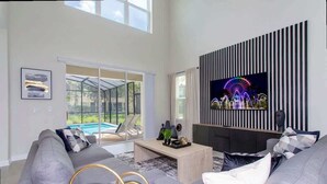 A modern living room with a large TV, grey sofas, a coffee table, and a glass door leading to a pool. - Family 5BD Spa Pool Game Loft Near Disney (Kissimmee)
