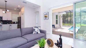 A modern living room with a grey sofa, a coffee table with a plant, and a view of a swimming pool through large glass doors. - Family 5BD Spa Pool Game Loft Near Disney (Kissimmee)