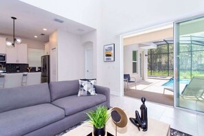 A modern living room with a grey sofa, a coffee table with a plant, and a view of a swimming pool through large glass doors. - Family 5BD Spa Pool Game Loft Near Disney (Kissimmee)