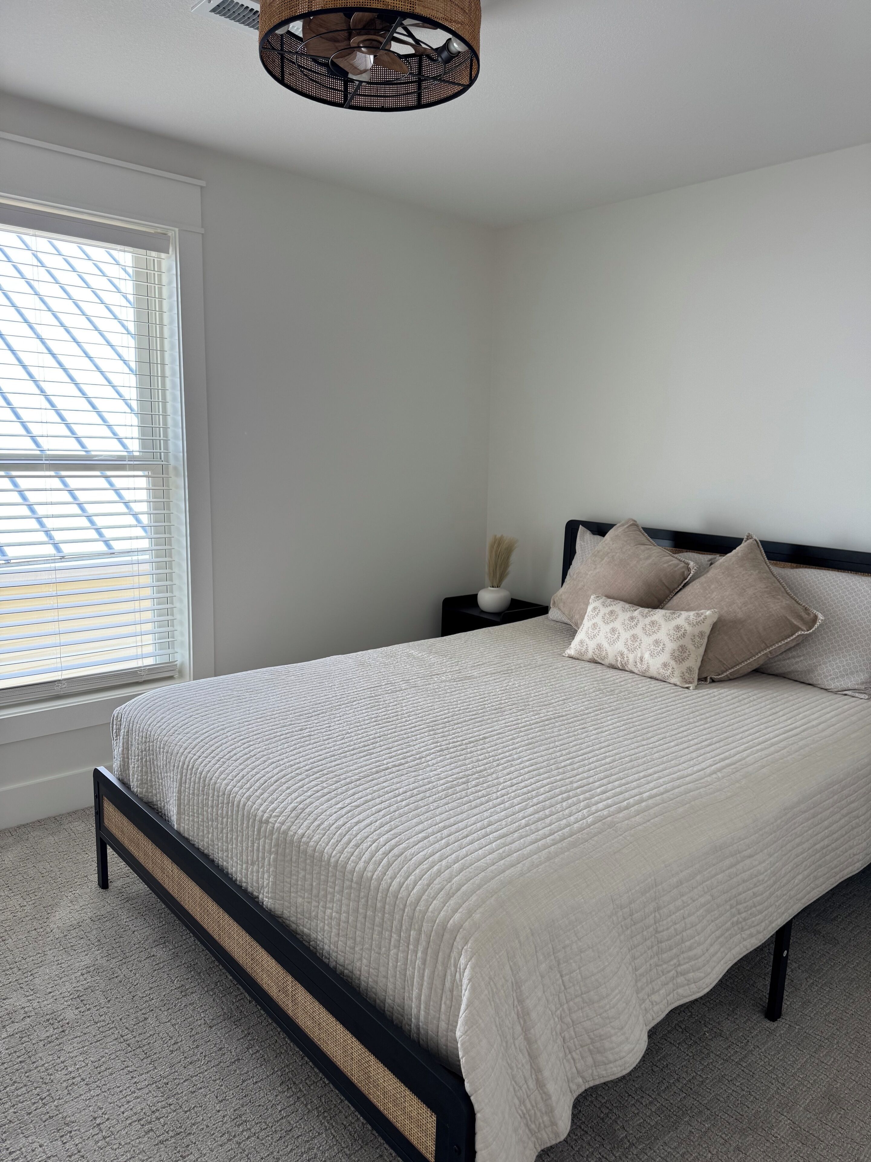 A neatly made bed with a white quilt, a black headboard, and several pillows. There is a small bedside table with a vase and a lamp. The room has white walls and a large window with blinds.