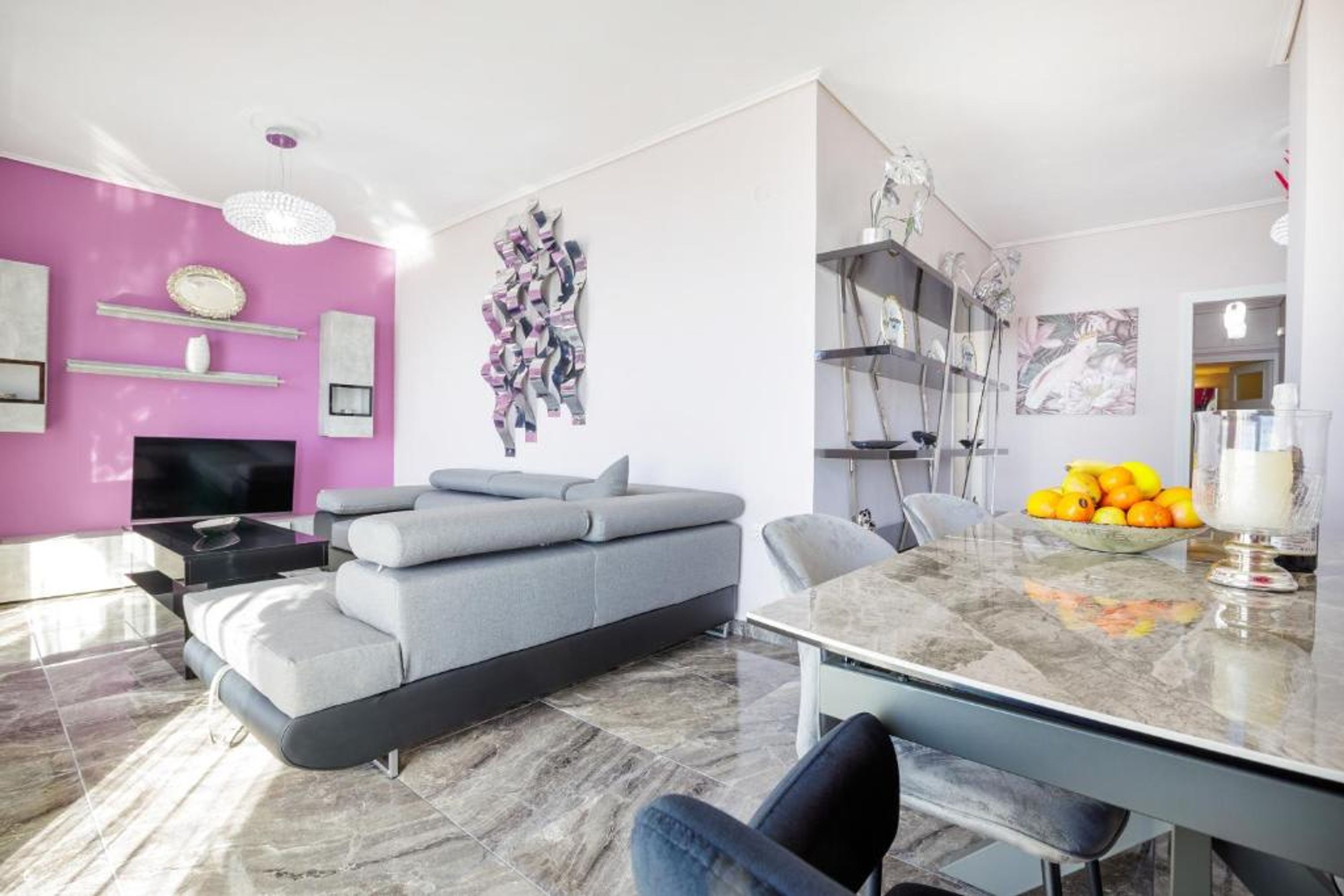 Uma sala de estar moderna com um sofá cinza, uma mesa de jantar com cadeiras, uma televisão e uma parede cor de rosa.