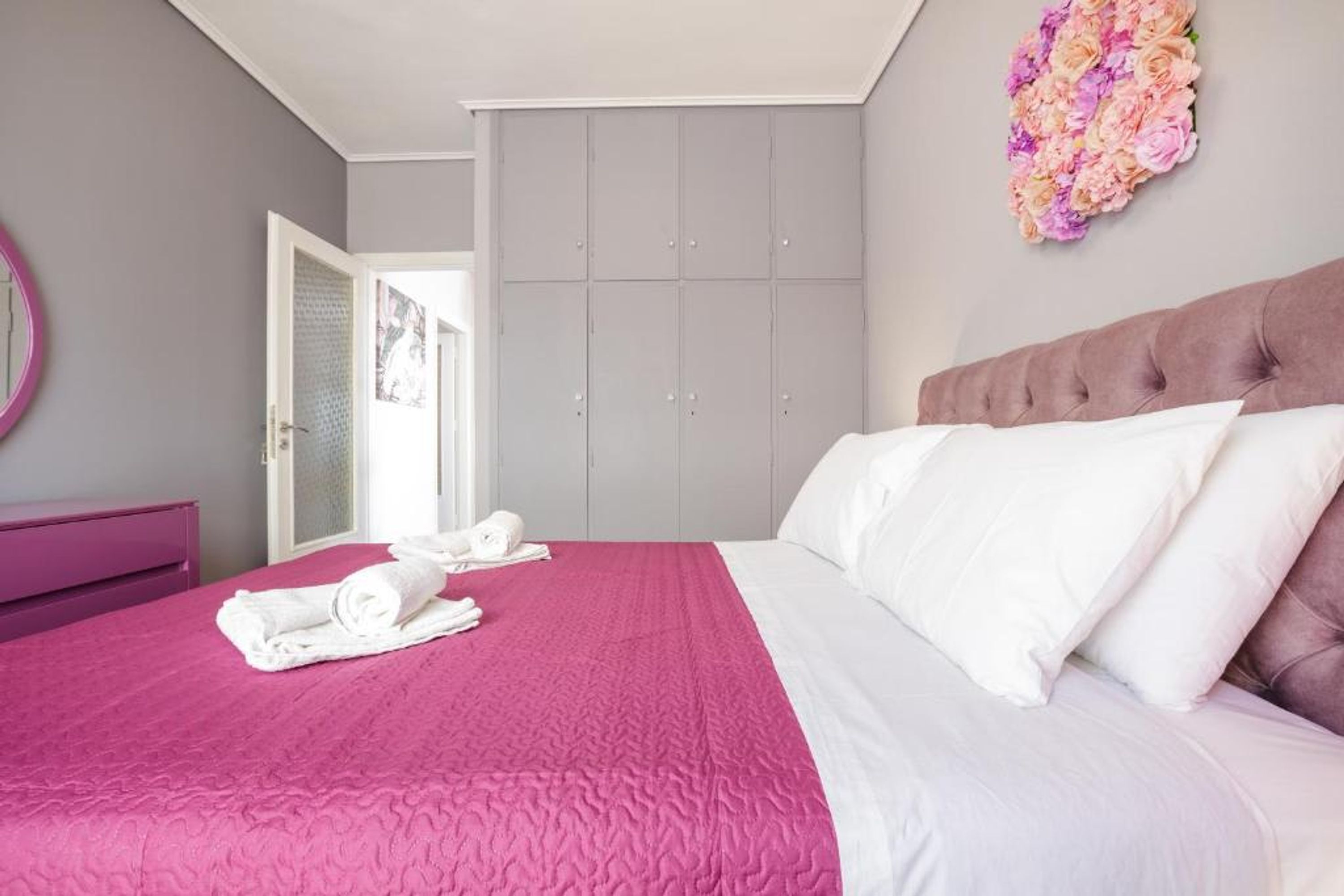 A bedroom with a pink bedspread, white pillows, a tufted headboard, a grey wardrobe, and a floral wall decoration.