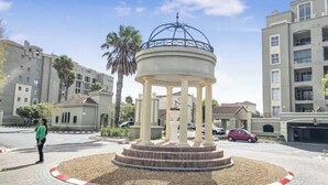 A gazebo with a dome roof, surrounded by a landscaped area with palm trees and a parking lot with cars. - 2 Bedroom Mountain View Penthouse Century City (Cape Town)
