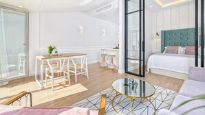 A modern hotel room with a glass door leading to a balcony, a bed with a teal headboard, a dining area with a wooden table and white chairs, and a small round table with a glass top. - Atico Sebastian Souviron - Penthouse in Malaga (Málaga)