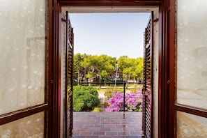 A view through a doorway with a brick floor and a metal railing, leading to a garden with trees and pink flowers.