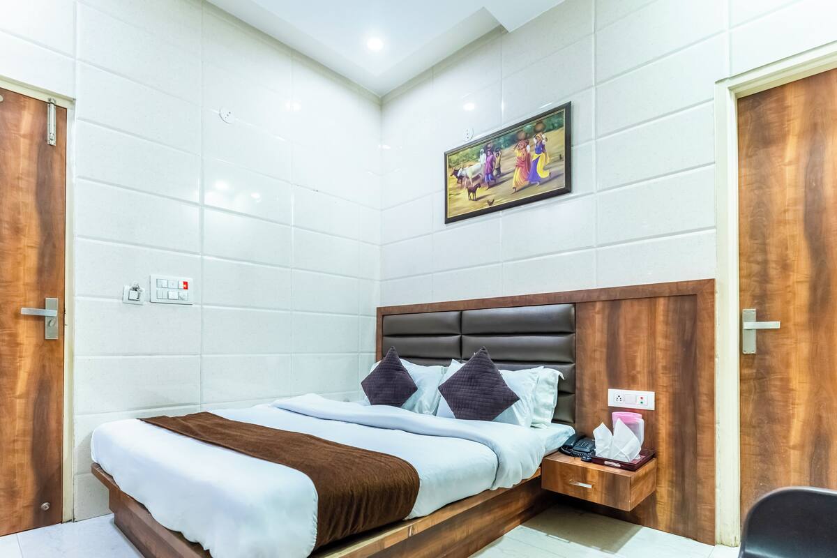 Une chambre d’hôtel avec un grand lit, une tête de lit en bois, des murs recouverts de carreaux blancs, un tableau accroché au mur et une porte en bois.