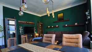 Un salon comprenant une table à manger en bois, deux canapés, un téléviseur et un mur vert agrémenté d’étagères.