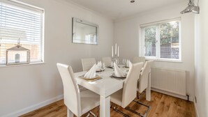 A dining area with a white table set for six, white chairs, a mirror on the wall, and a window with blinds. - Spacious 6 Bedroom House in Maidenhead/ Windsor (Maidenhead)