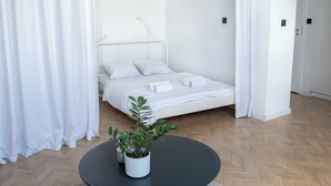 Een wit bed met witte lakens, een potplant op een ronde tafel en een houten vloer.