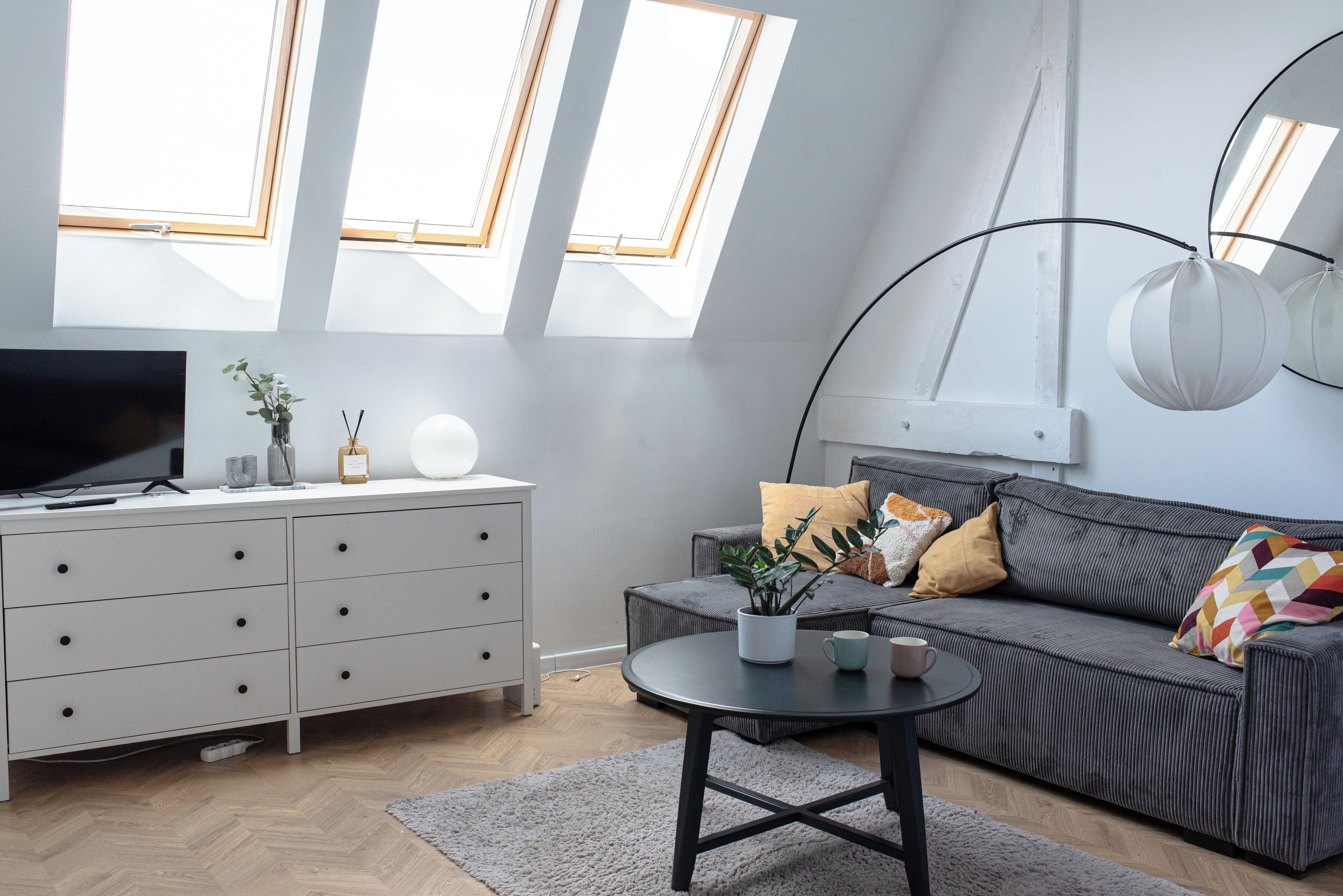 A modern living room with a grey sofa, a round coffee table, and a TV on a white dresser. There are two skylights and a hanging light fixture.