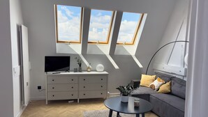 Een moderne woonkamer met een televisie, bank en een klein bijzettafeltje. Boven de bank bevinden zich dakramen en er staat een witte dressoir met lades.