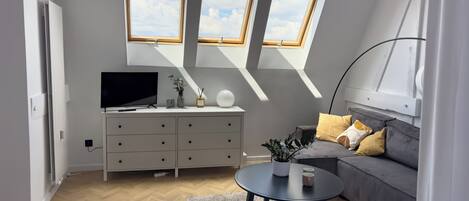 Una sala de estar moderna con televisor, sofá y una pequeña mesa de centro. Hay tragaluces sobre el sofá y un aparador blanco con cajones.