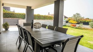 A covered outdoor dining area with a rectangular table and chairs, a decorative centerpiece, and a view of a garden and neighboring buildings. - Holiday home Galižana (Galižana)