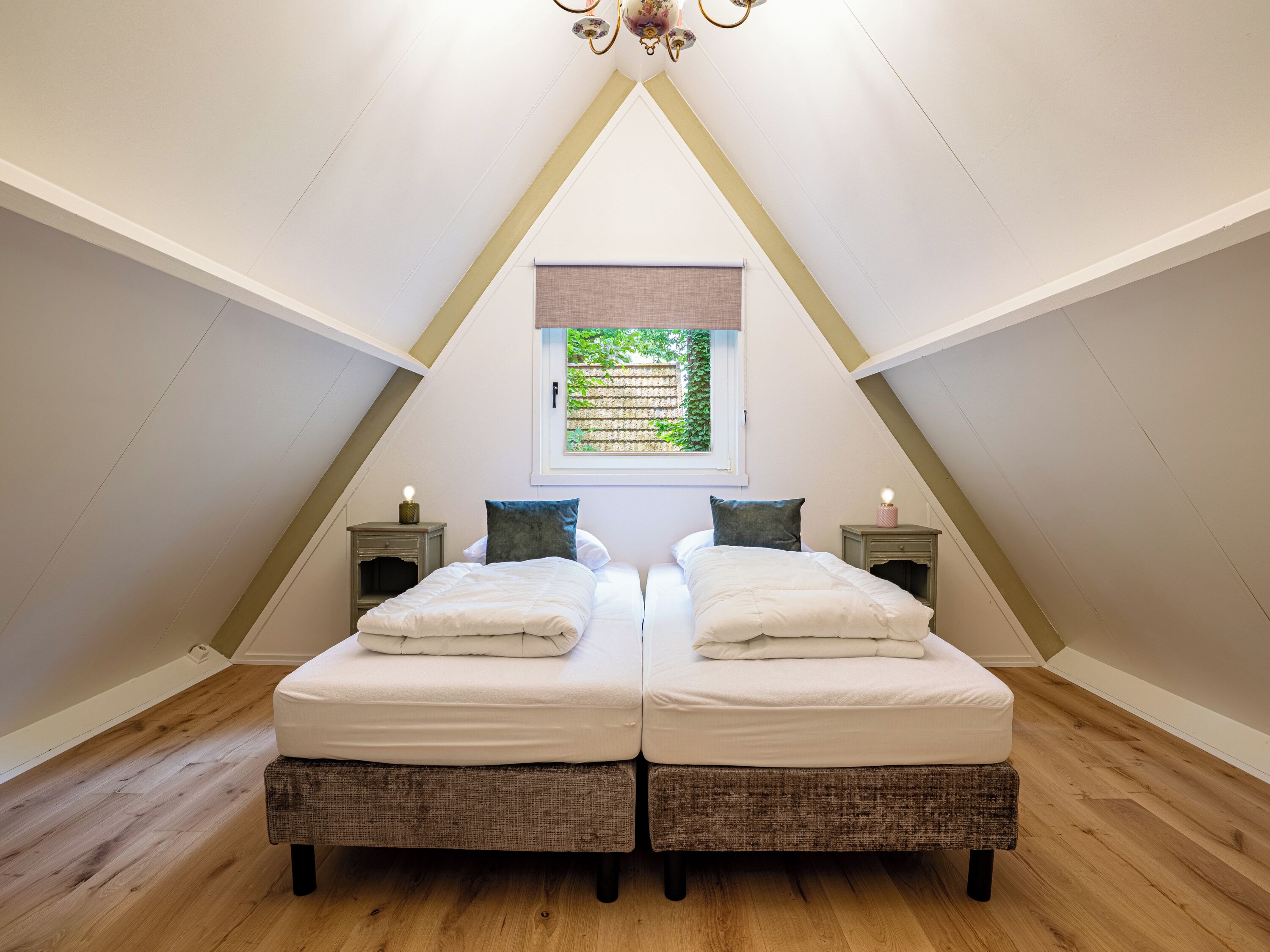 Quarto com duas camas, piso de madeira, janela com vista para área verde e um lustre.