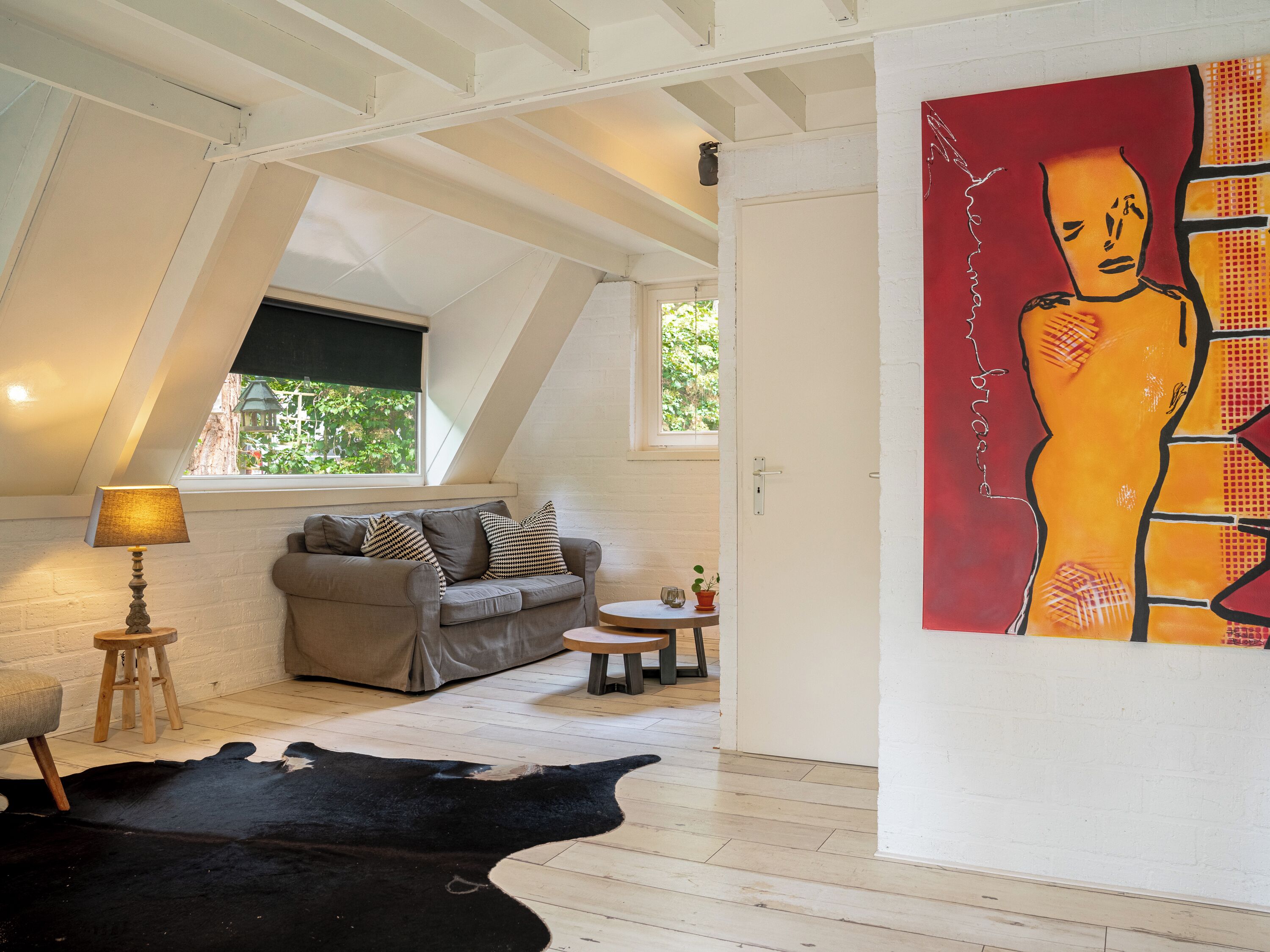 A cozy living room with a sloped ceiling, a grey sofa, a small round table, and a painting on the wall.